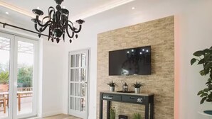A modern living room with a chandelier, a flat-screen TV mounted on a textured wall, a console table with decorative items, and a sliding glass door leading to an outdoor area. - Deluxe Apartment Marghareta (Dubrovnik)