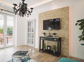 A modern living room with a chandelier, a flat-screen TV mounted on a textured wall, a console table with decorative items, and a sliding glass door leading to an outdoor area. - Deluxe Apartment Marghareta (Dubrovnik)
