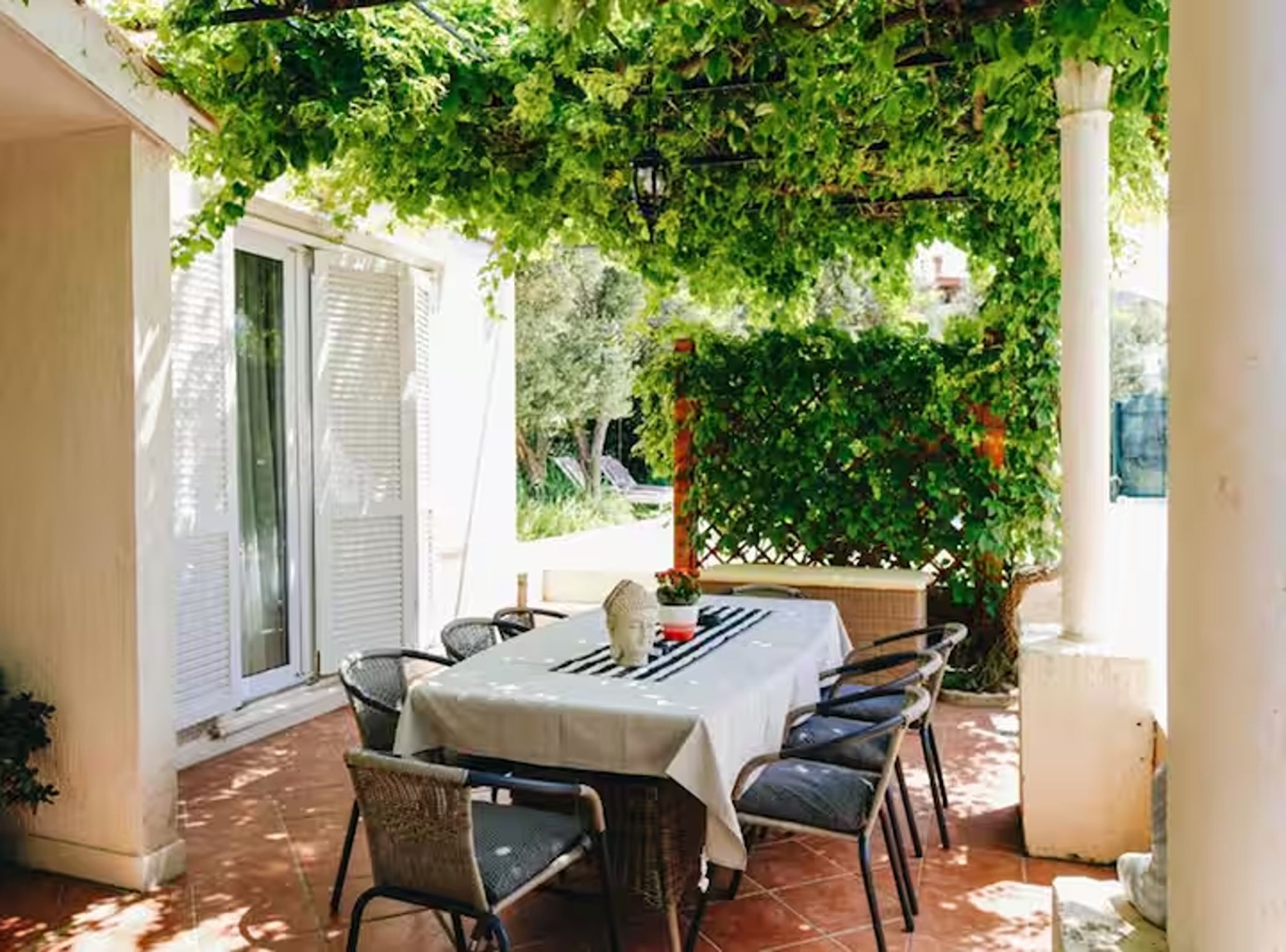 An outdoor patio with a table and chairs, surrounded by greenery and a white column.