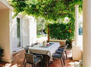 An outdoor patio with a table and chairs, surrounded by greenery and a white column. - Deluxe Apartment Marghareta (Dubrovnik)