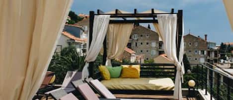 Un balcon doté d’un lit à baldaquin, de chaises longues et offrant une vue sur les immeubles.