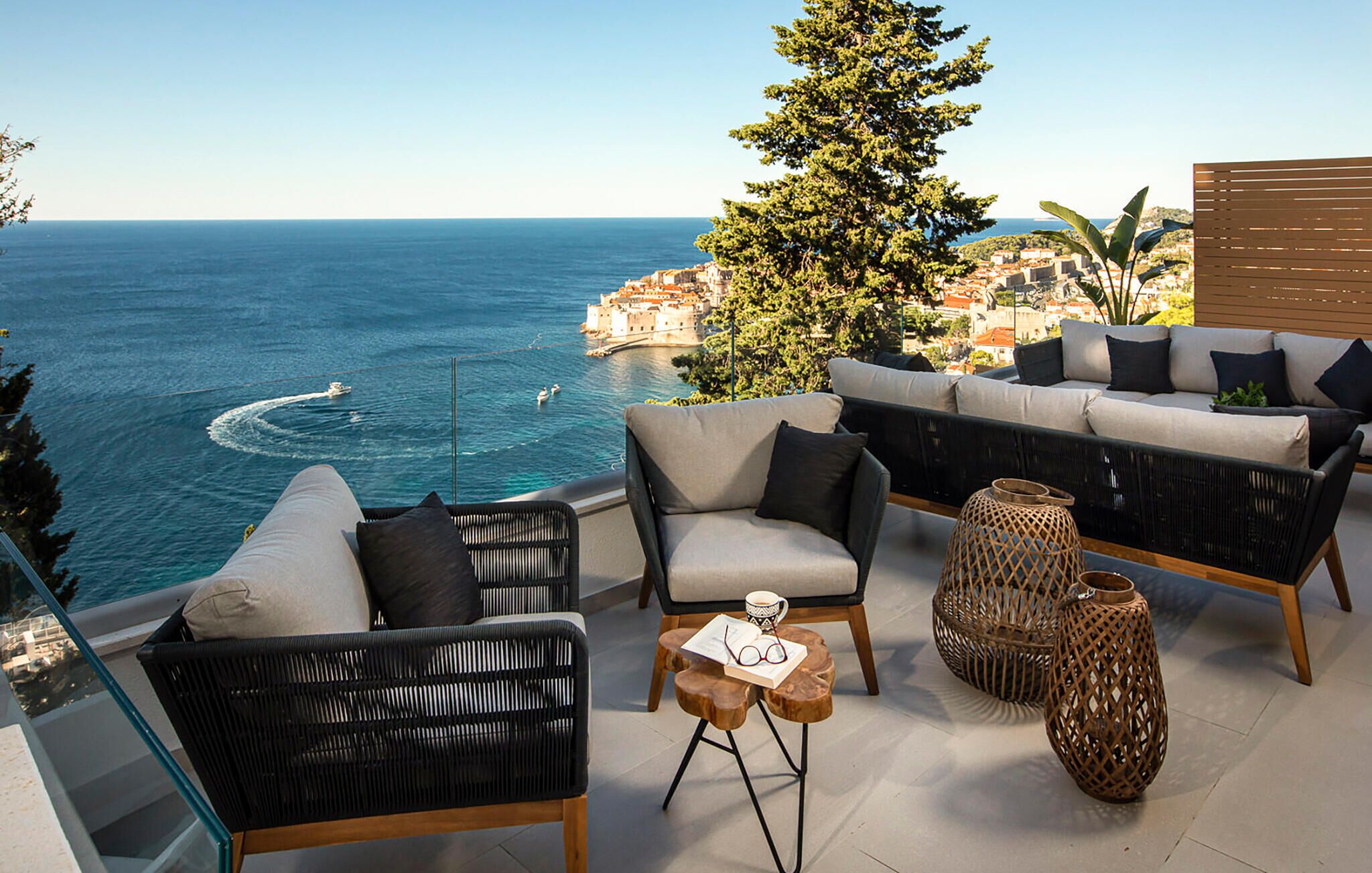 A terrace with wicker furniture, a small table with a book and glasses, and a view of the sea and a historic building.