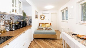 A compact room with a kitchenette, a bed with pillows, a round mirror, and a window with blinds. - Marghareta Nest 2 - TOP Location (Dubrovnik)