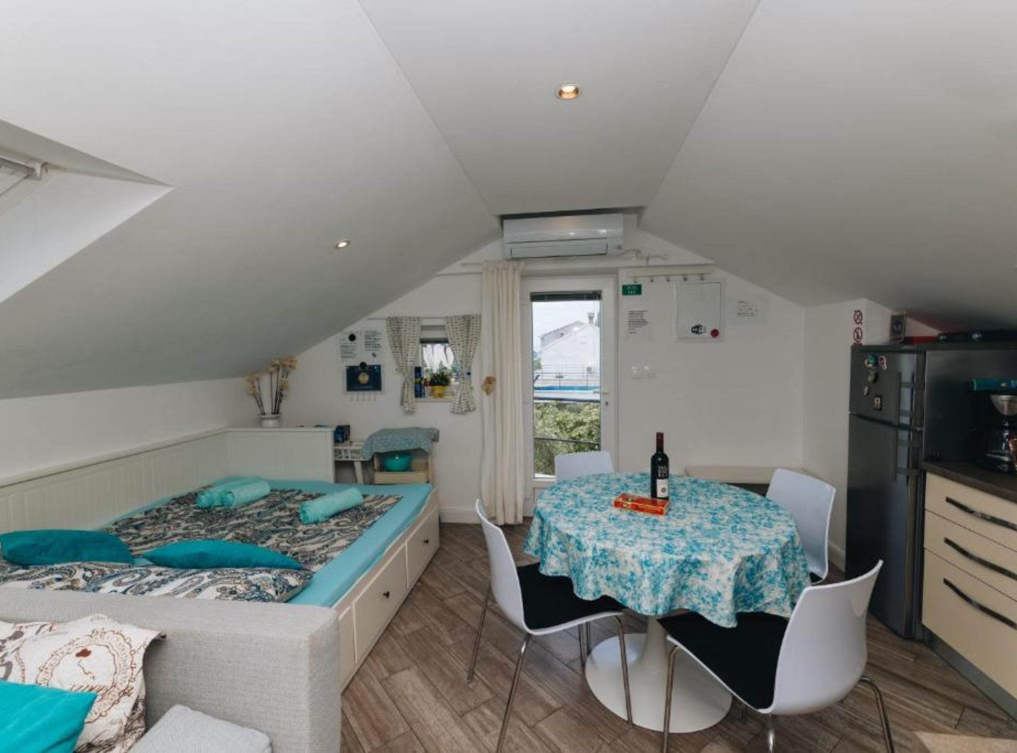 Una habitación compacta con cama, una mesa de comedor, sillas, kitchenette y un ventanal.