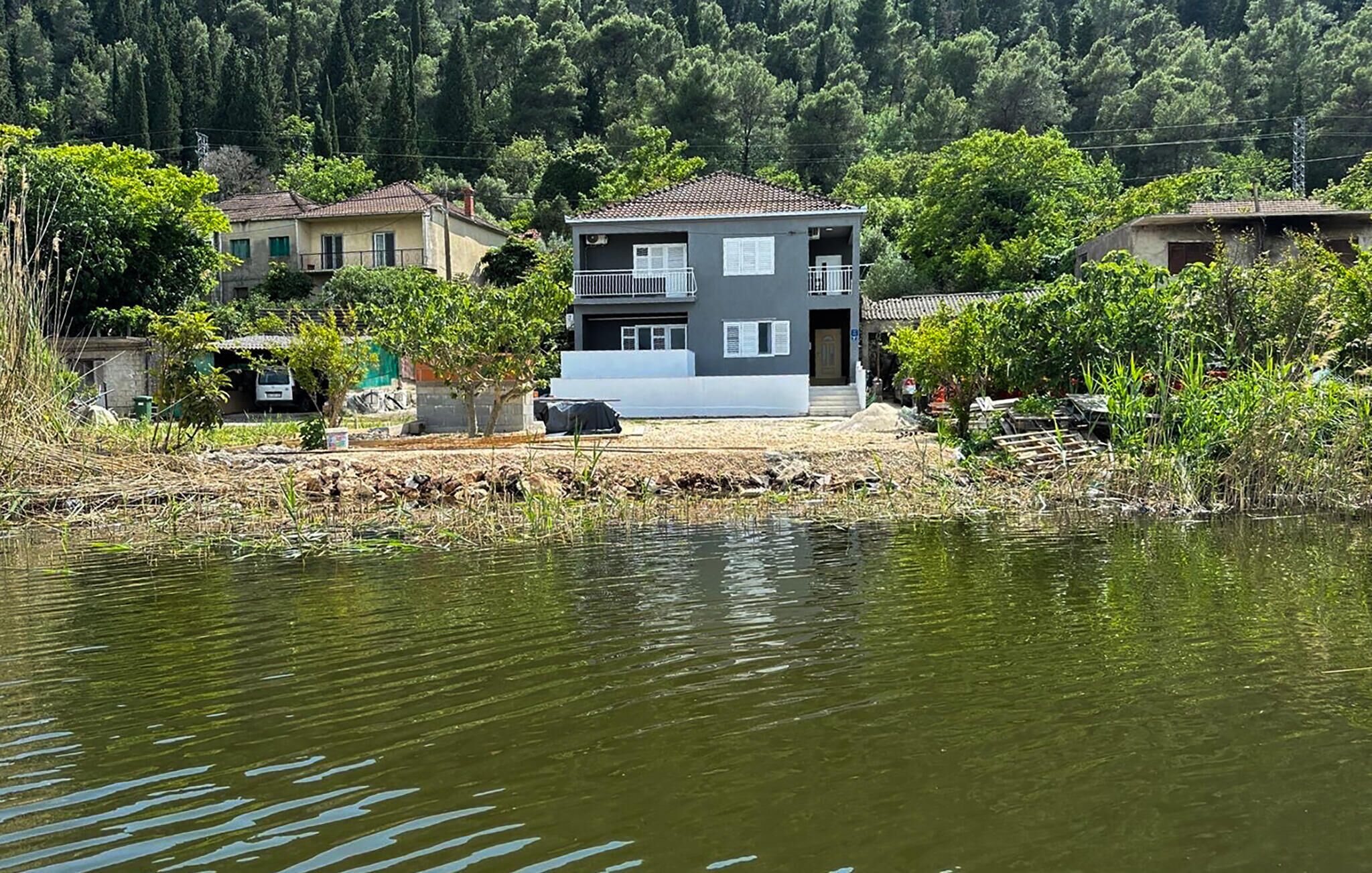 Une maison à deux étages avec un balcon, située au bord d’un plan d’eau et entourée de verdure.