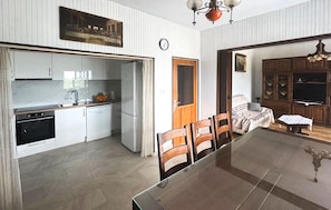Uma cozinha moderna com armários brancos, uma área de jantar com mesa de vidro e uma sala de estar com sofá e aparador de madeira.