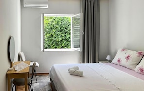 Een slaapkamer met een bed, een bureau, een stoel, een raam met luiken en uitzicht op groen.