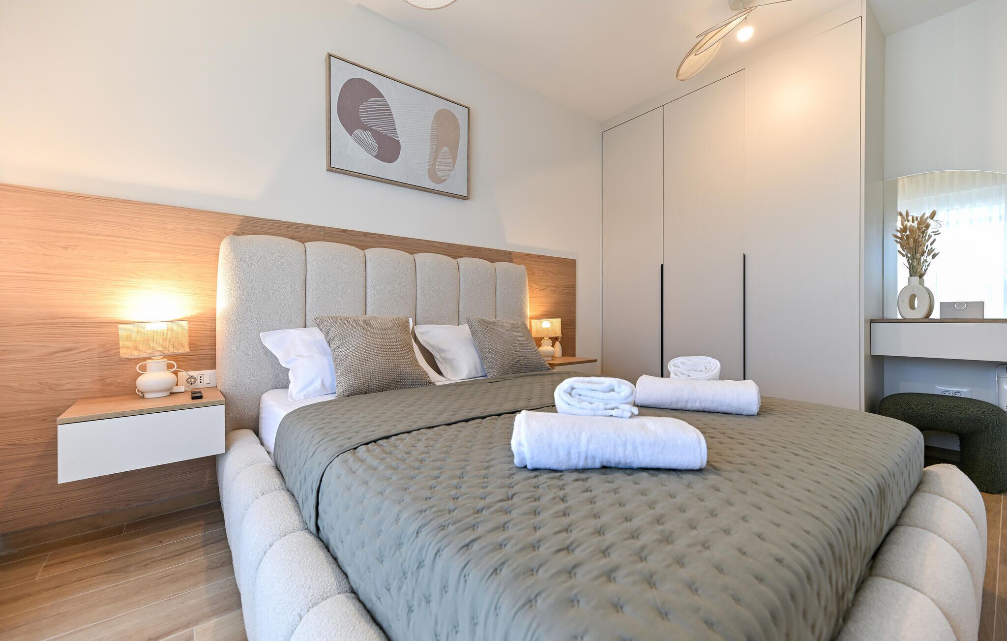 A modern hotel room with a large bed, a bedside table with a lamp, a wall-mounted artwork, and a view of the city.