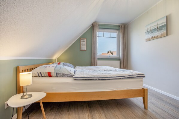 A bedroom with a single bed, a bedside table with a lamp, a window with curtains, and a painting on the wall.