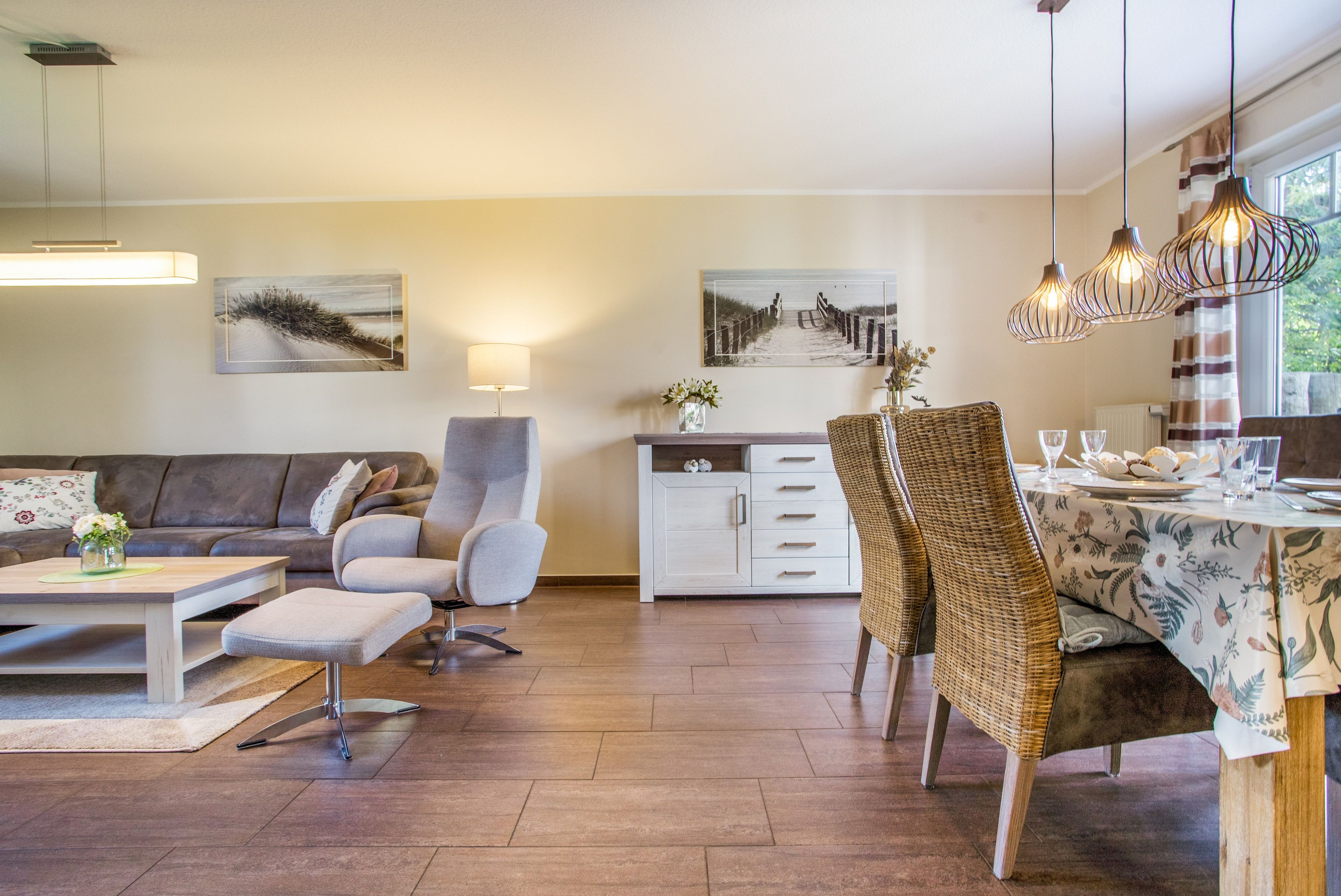 A modern living room with a sofa, armchair, and dining table. There are wall-mounted artworks and a window with curtains.