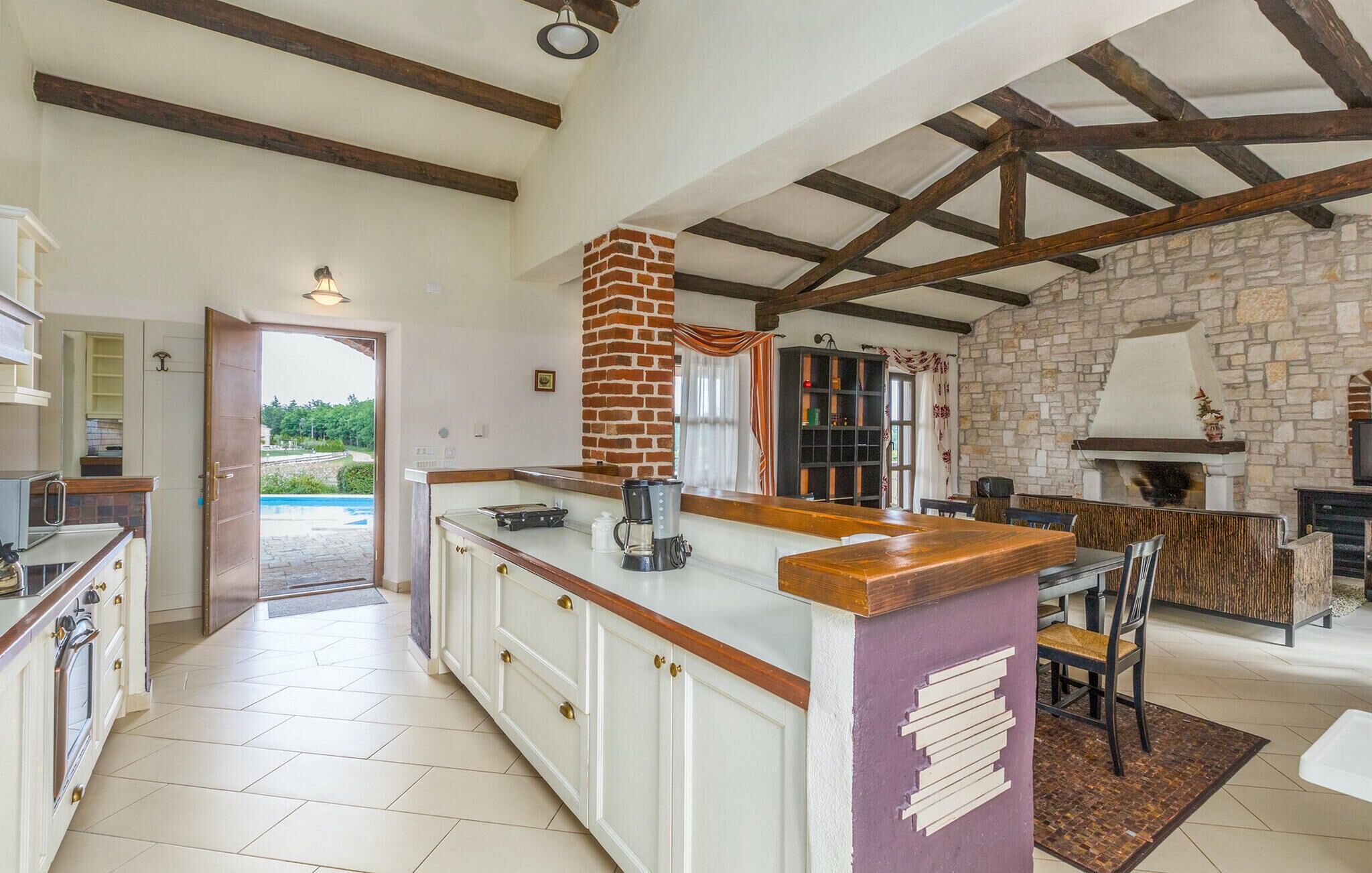 A spacious kitchen with a large island, exposed wooden beams, stone walls, and a view of a swimming pool.