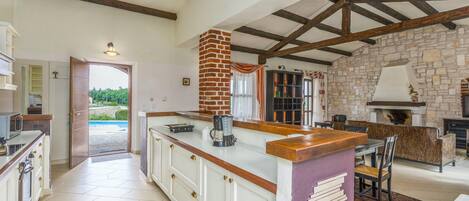 A spacious kitchen with a large island, exposed wooden beams, stone walls, and a view of a swimming pool.