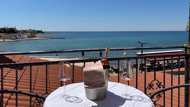 Une terrasse avec une table, une bouteille et un verre, donnant sur une étendue d’eau et des bâtiments.