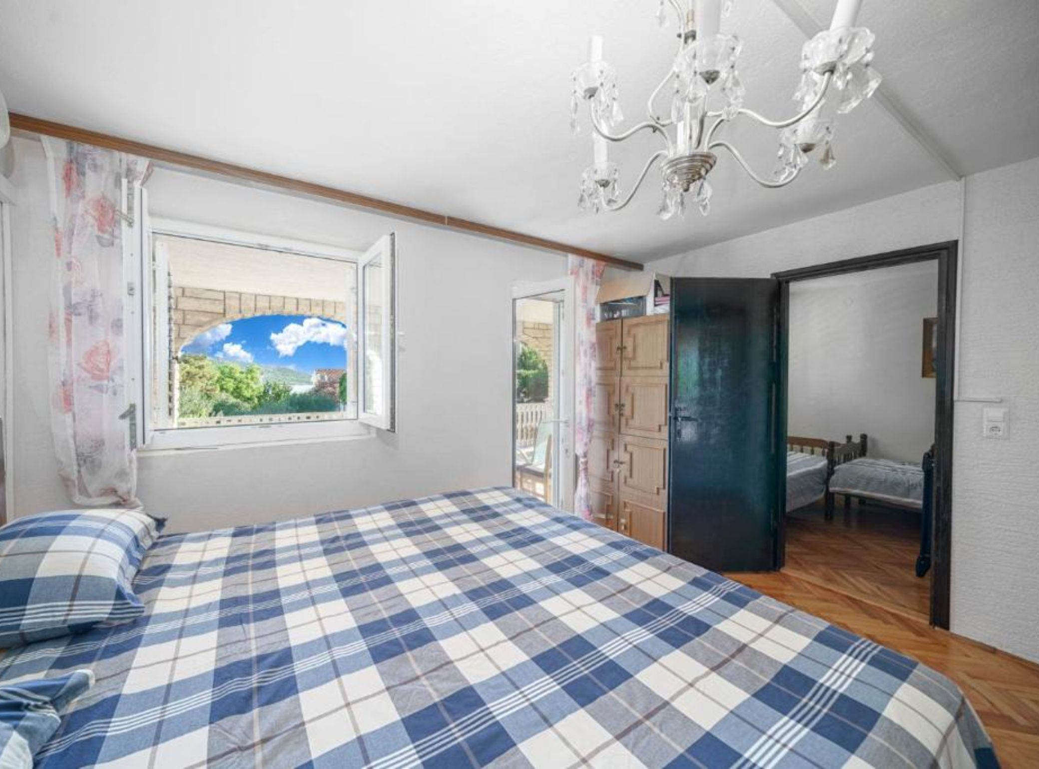 A bedroom with a checkered bedspread, a chandelier, a window with a view, and a door leading to another room.