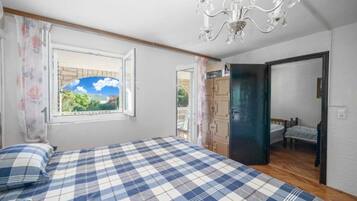 A bedroom with a checkered bedspread, a chandelier, a window with a view, and a door leading to another room.