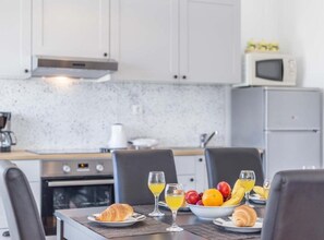 A modern kitchen with a dining table set for breakfast, including croissants, fruit, and juice glasses.