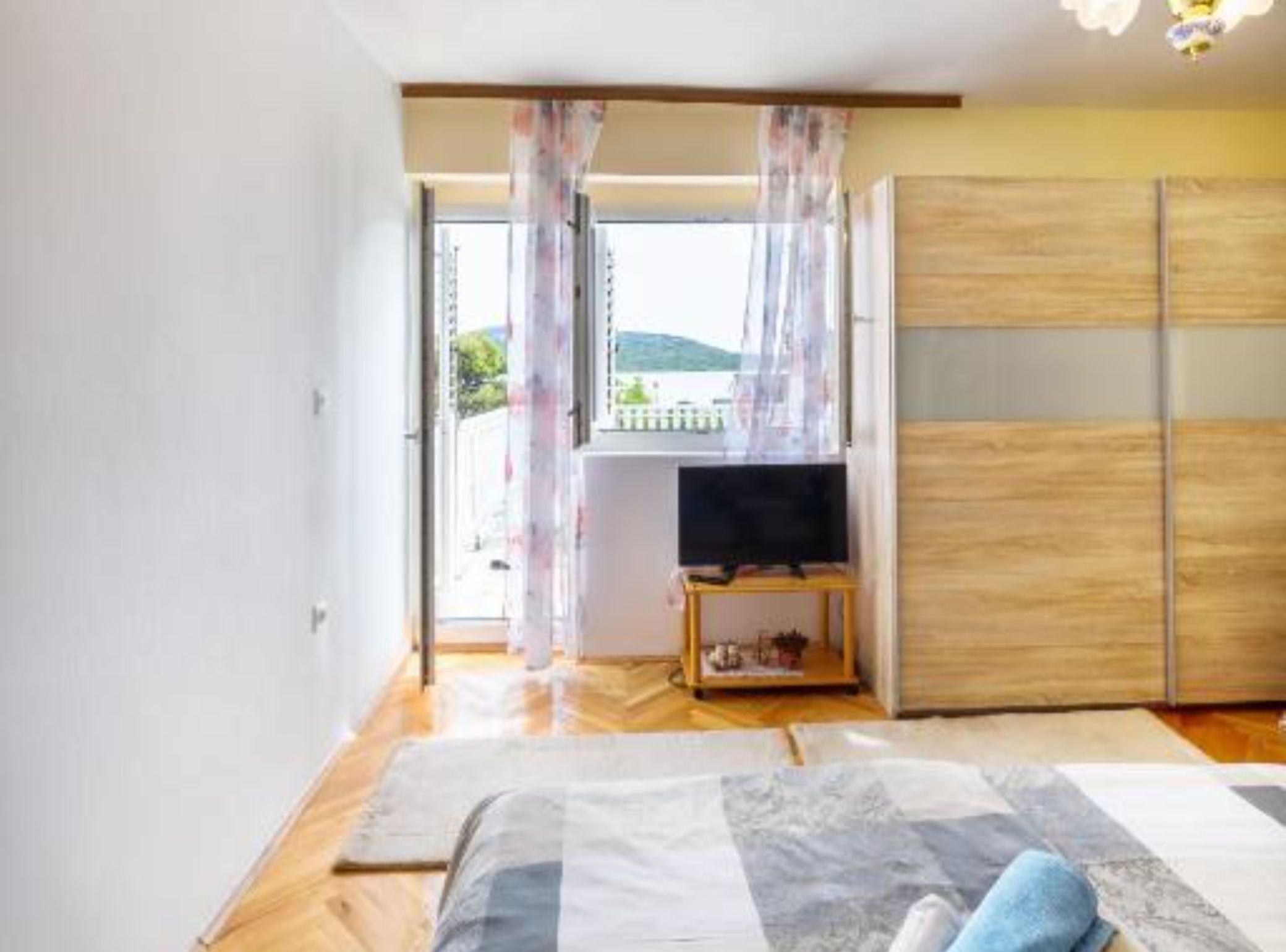 A bedroom with a bed, a TV on a stand, a wooden wardrobe, and a view of the outdoors.