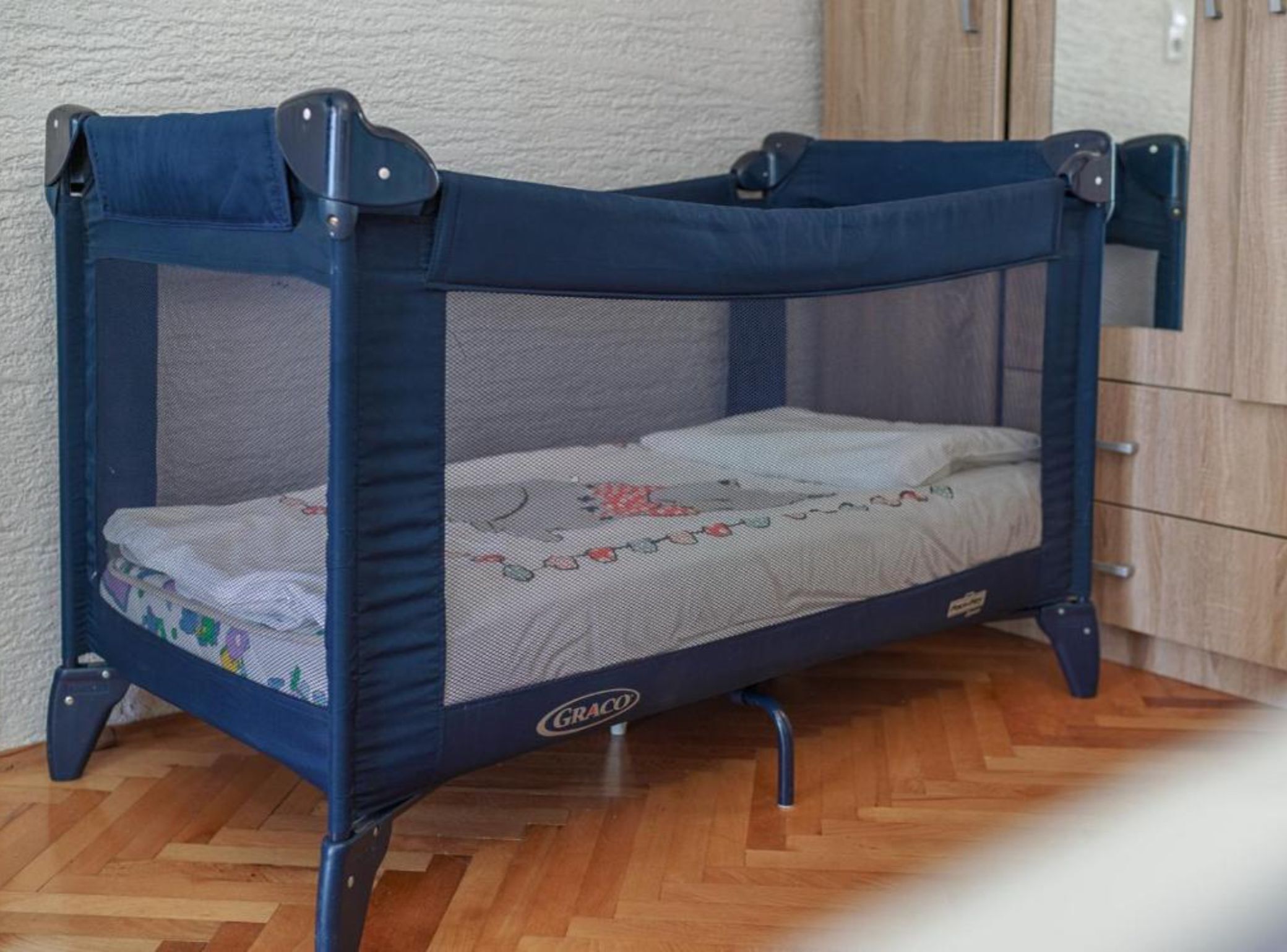 A blue Graco baby playpen with a mesh top and a mattress inside, placed on a wooden floor.
