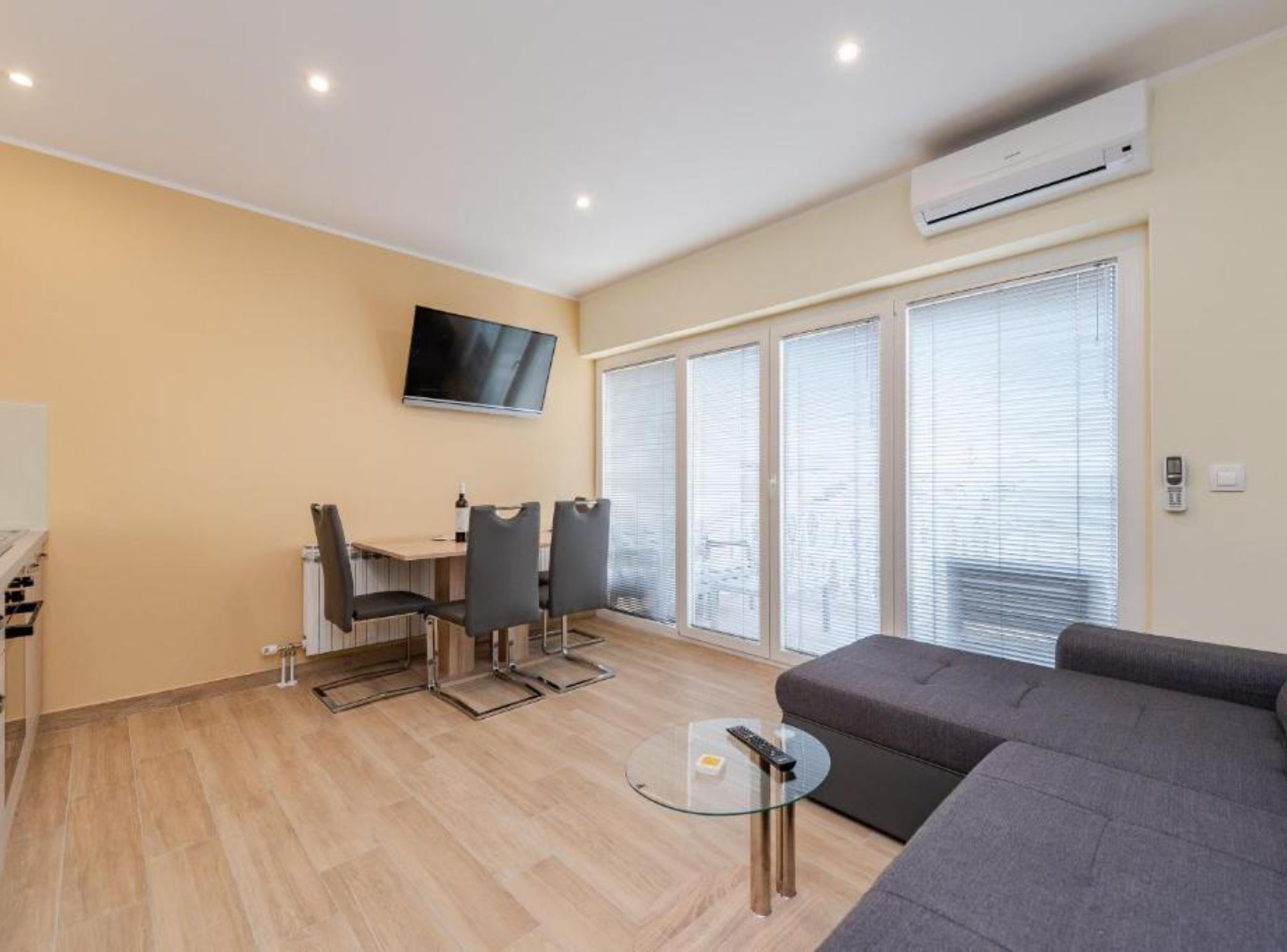Una sala de estar moderna con un sofá, una mesa de centro de vidrio, un televisor montado en la pared y una zona de comedor con sillas y una mesa.