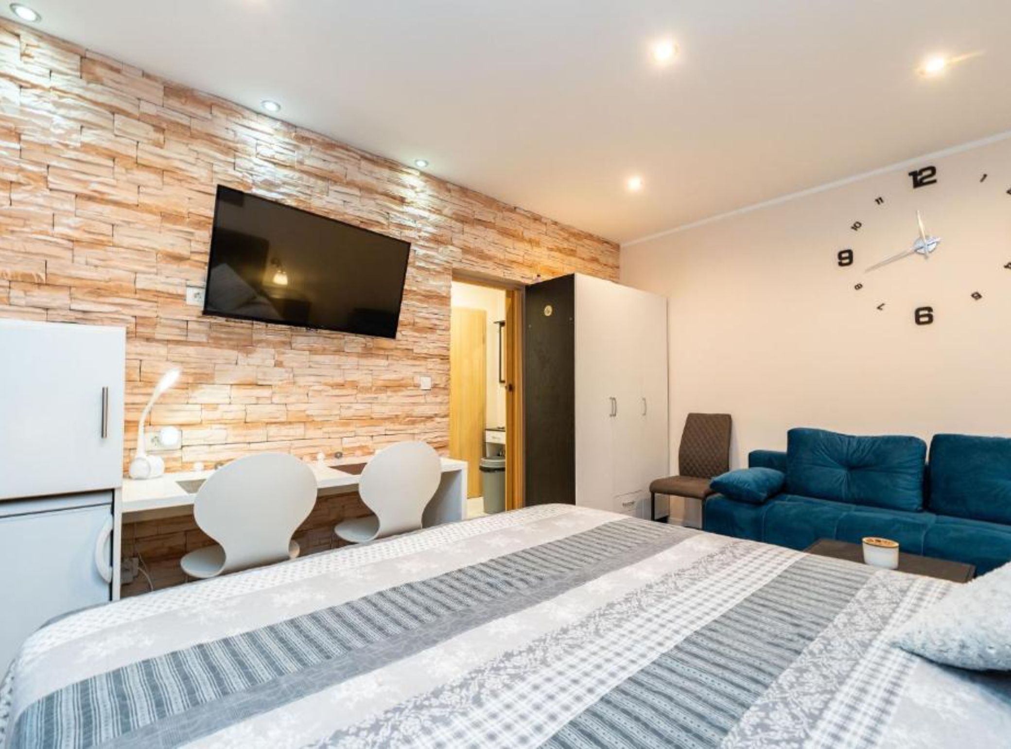 Ein modernes Hotelzimmer mit Backsteinwand, Flachbildfernseher, einem Bett mit gemusterten Bettwäsche, einem blauen Sofa und einem Schreibtisch mit Stühlen.