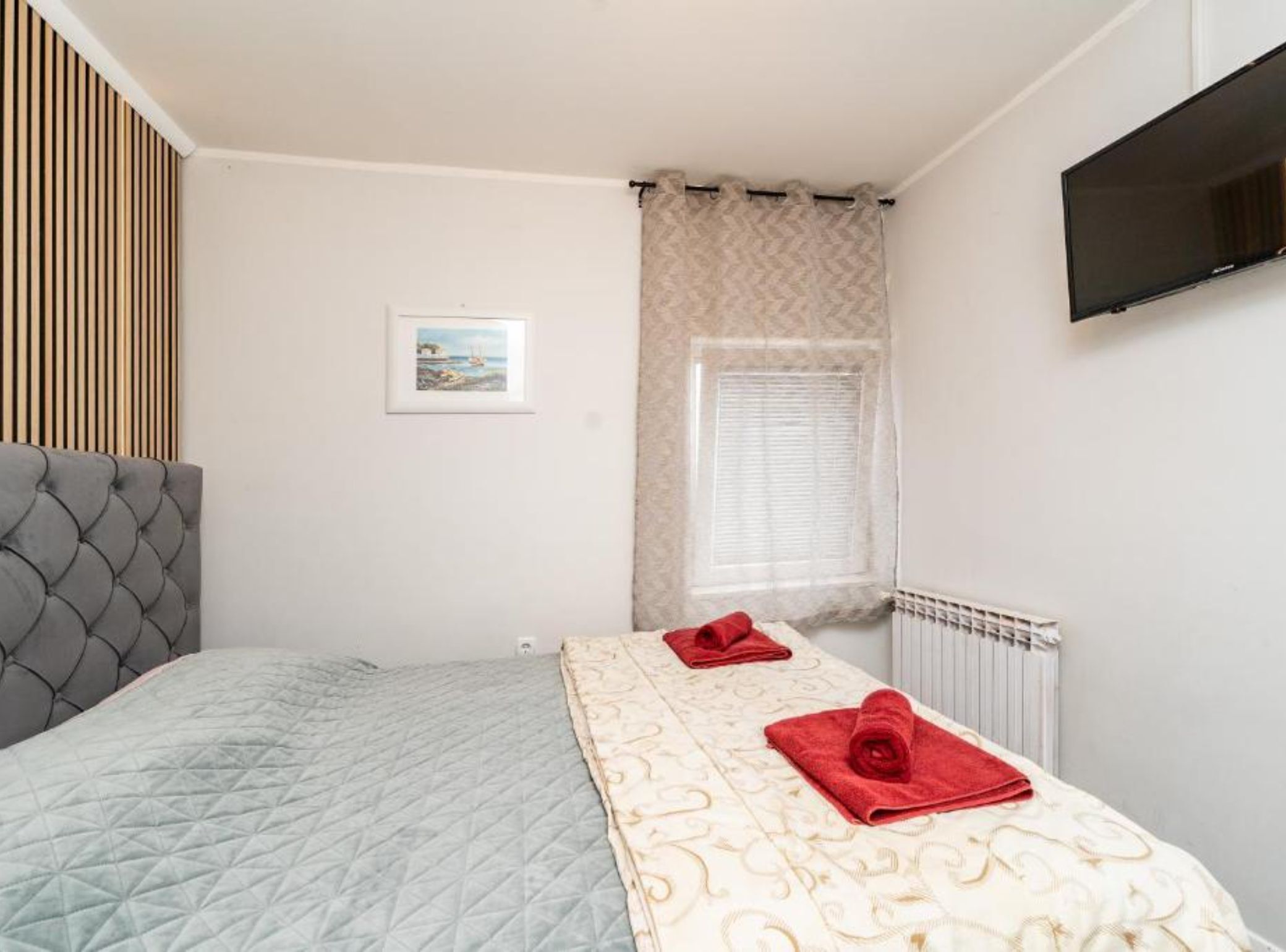 A bedroom with a bed, a window with curtains, a radiator, and a flat-screen TV mounted on the wall.