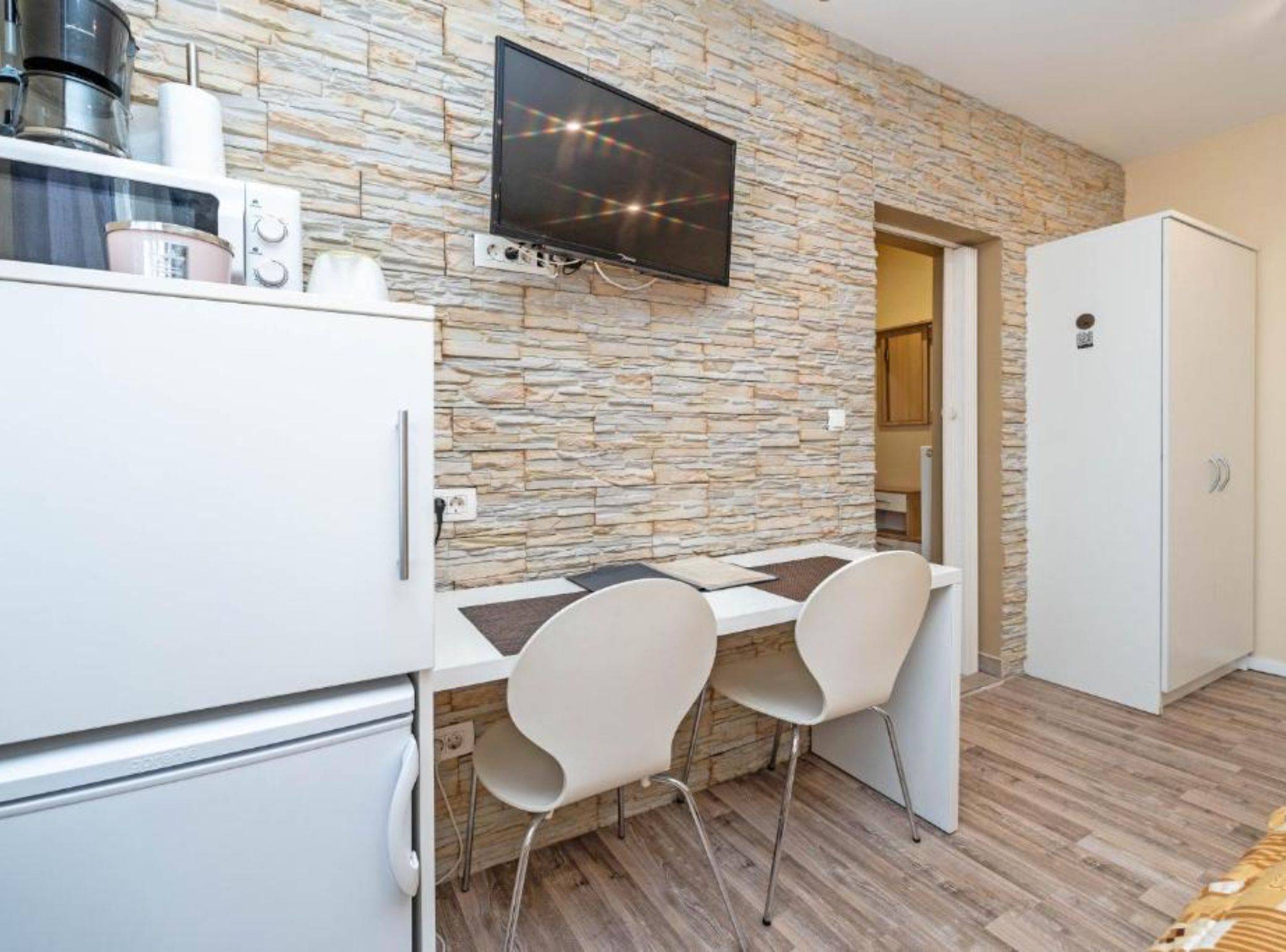 Een kleine keuken met een aanrecht van steen, een flatscreen televisie, een koelkast en twee witte stoelen.