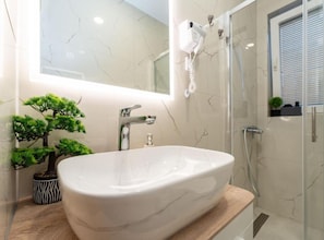 A bathroom with a white sink, a hairdryer mounted on the wall, a glass shower enclosure, and a small potted plant. - Bračić Premium Apartments With Outdoor Pool and Sauna (Sukosan)