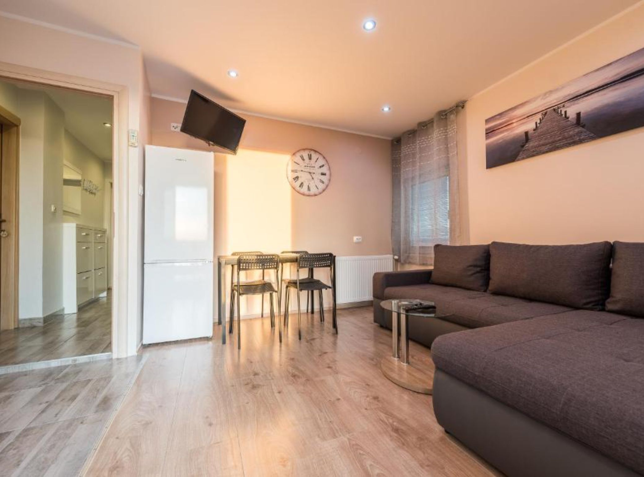 Una sala de estar con un sofá, una mesa de comedor con sillas, un televisor de pantalla plana montado en la pared y un reloj.