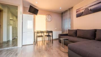 Una sala de estar con un sofá, una mesa de comedor con sillas, un televisor de pantalla plana montado en la pared y un reloj.