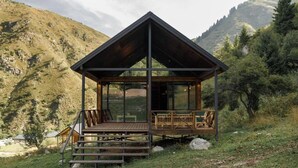 Una cabaña de madera con cerramiento de vidrio, terraza de madera y escalones que conducen a ella, con montañas al fondo.