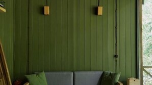 Una habitación con piso de madera, pared verde, sofá de madera con cojines y ventana con vista a follaje.