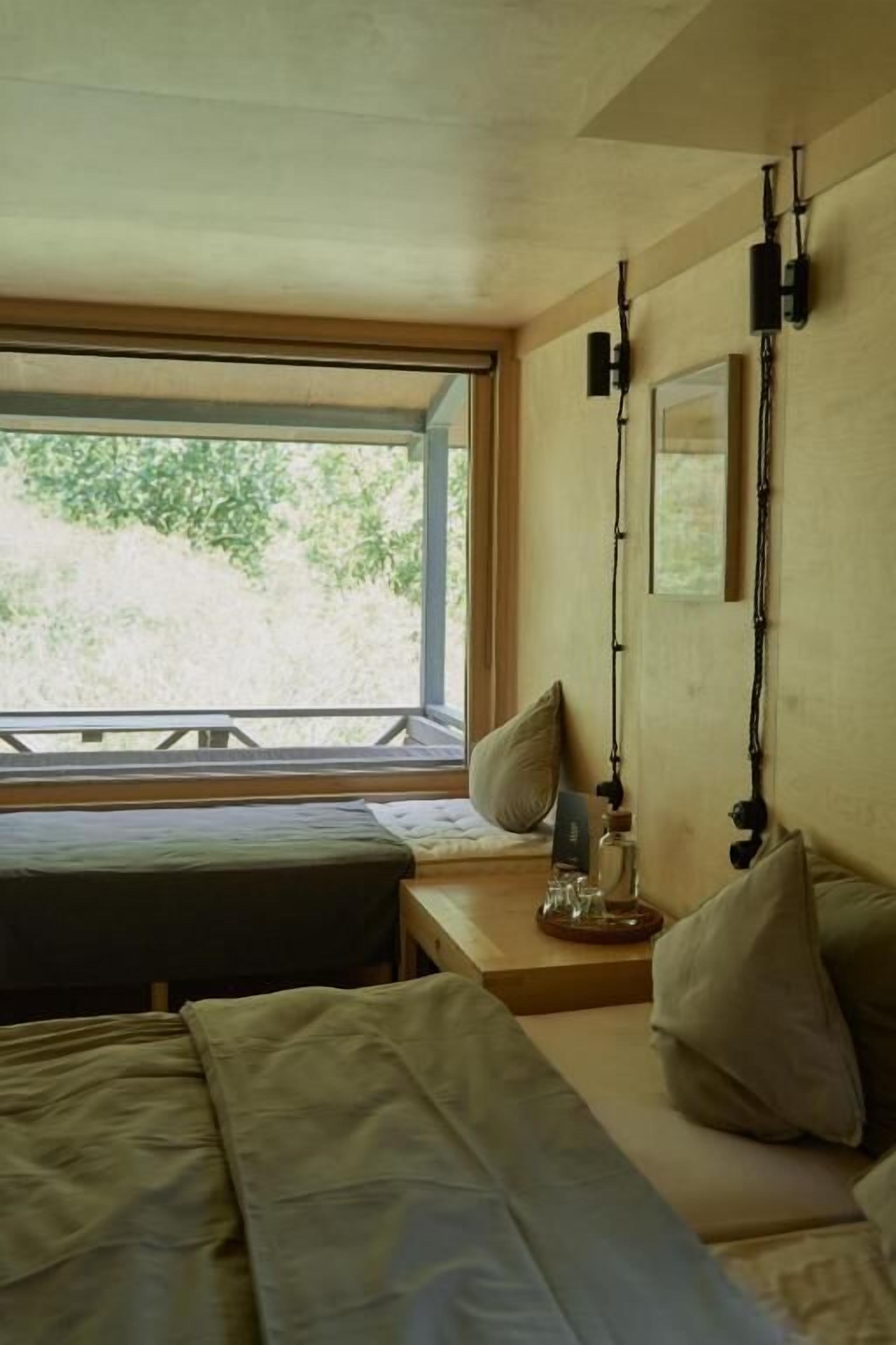 Ein Zimmer mit einem Bett, einem kleinen Tisch, einer Lampe und einem Fenster mit Blick auf Grünflächen.
