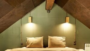 Un dormitorio con techo inclinado, una cama con ropa de cama color beige, dos luces fijadas a la pared y un radiador blanco.