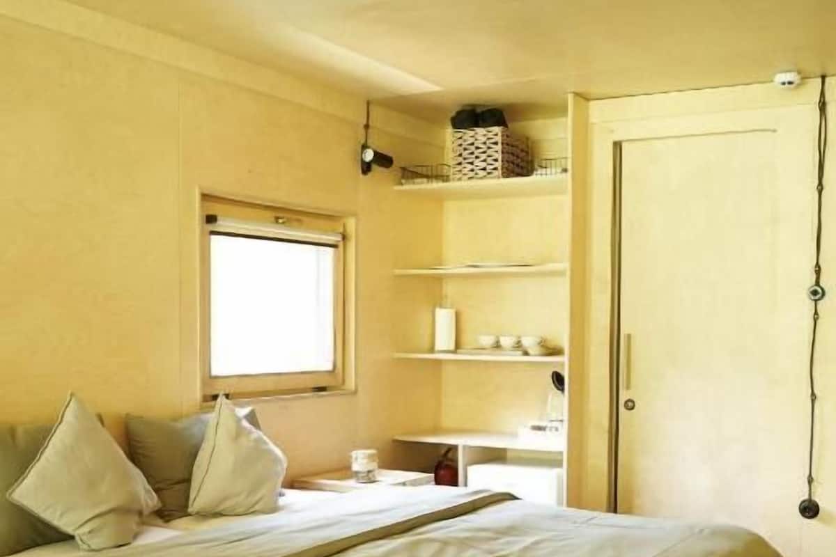 A single bed with a wooden frame, a bedside table, a small window, and a door with a hanging light fixture.