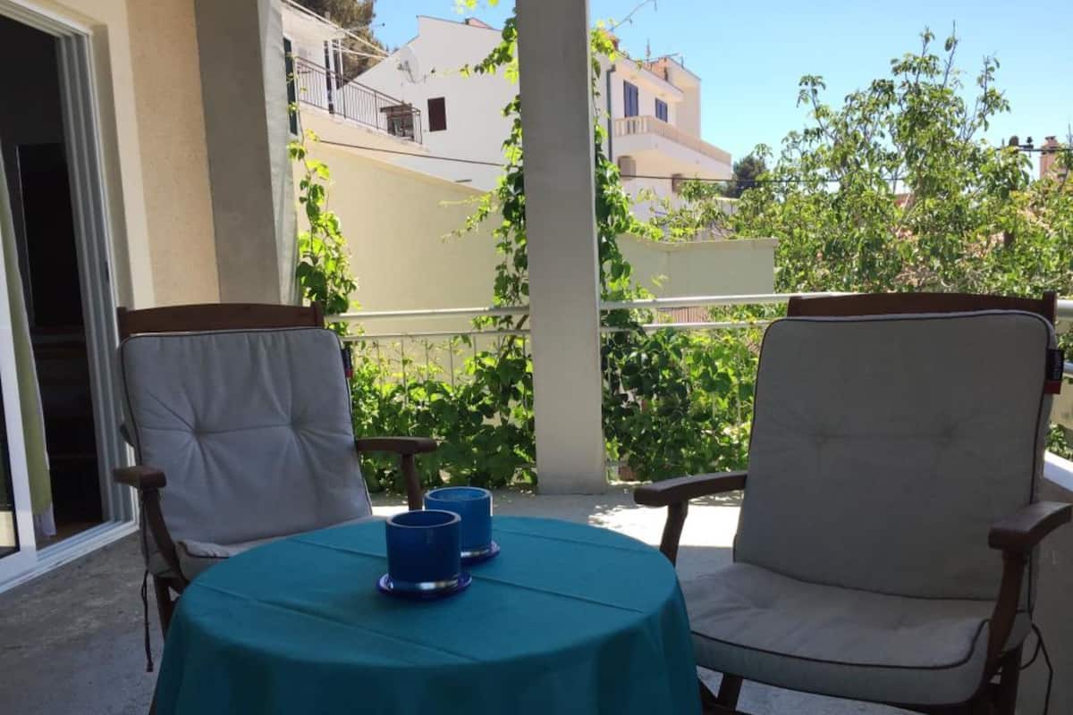 Un balcón con dos sillas y una mesa, cubierta con un mantel verde, y dos recipientes azules sobre la mesa.