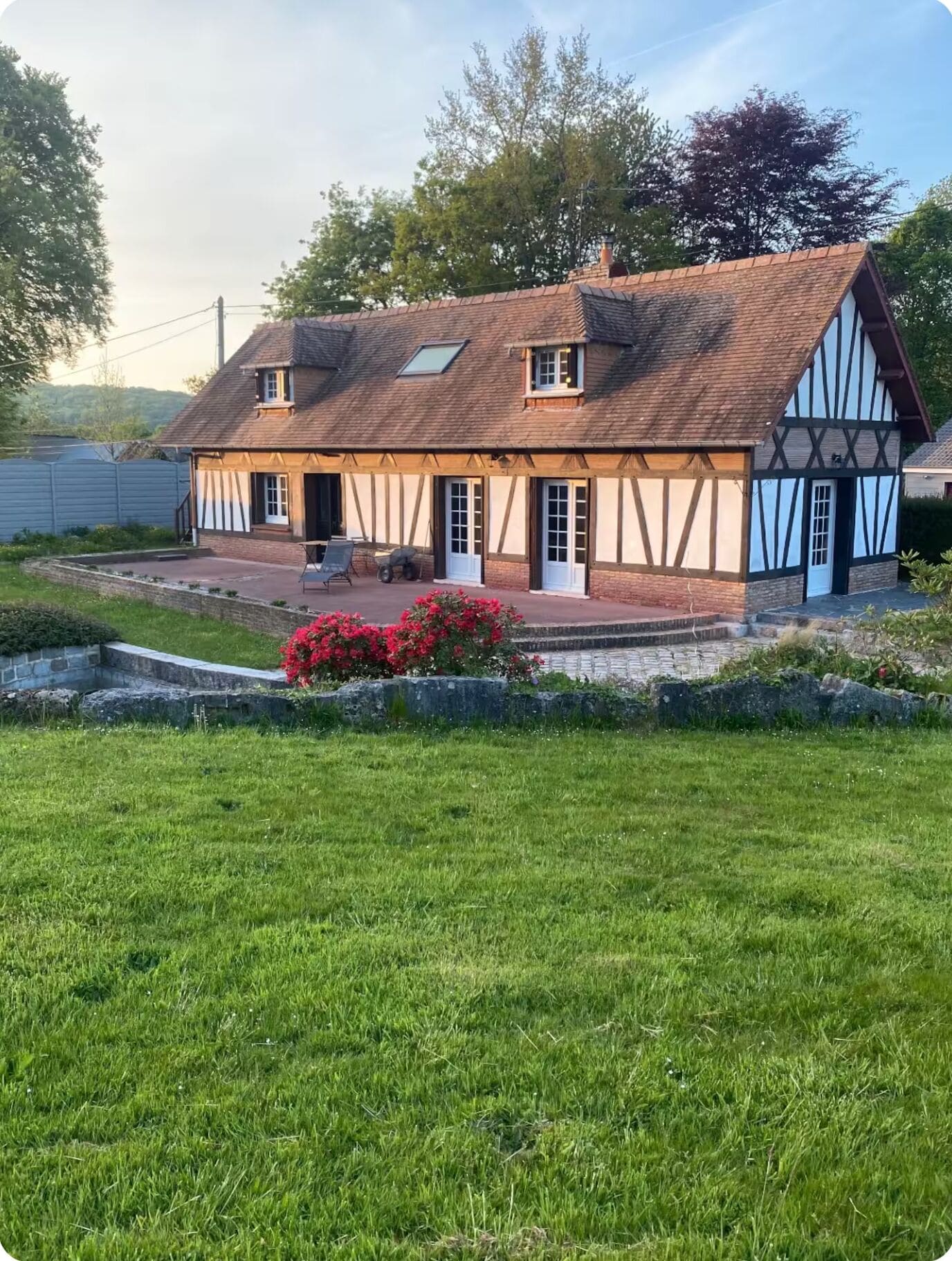 Una casa tradicional de entramado de madera con techo inclinado, un porche delantero y un pequeño jardín con flores rojas.