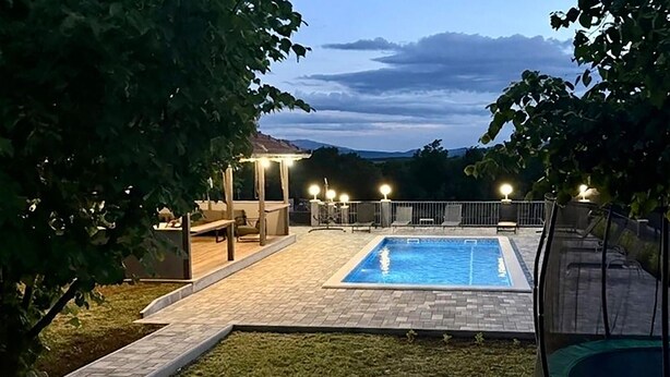 Patio trasero con piscina, un trampolín y un área de estar techada con luces.