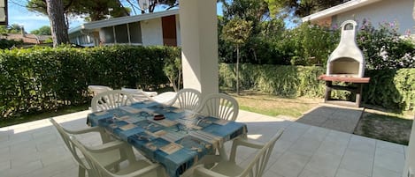 An outdoor patio with a table and chairs, a barbecue grill, and a view of the surrounding garden and trees.