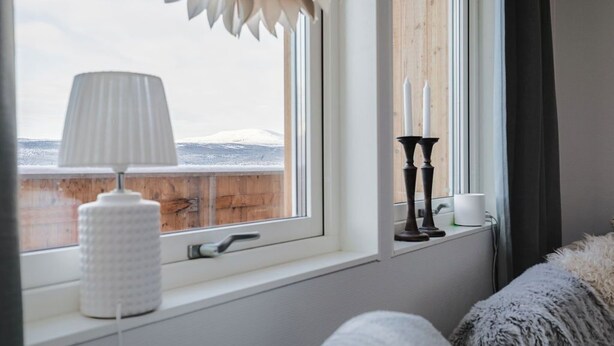 A room with a window, a white lamp, a candle holder, and a view of a snowy landscape. - Olen Village - Funäsdalen (Funäsdalen)