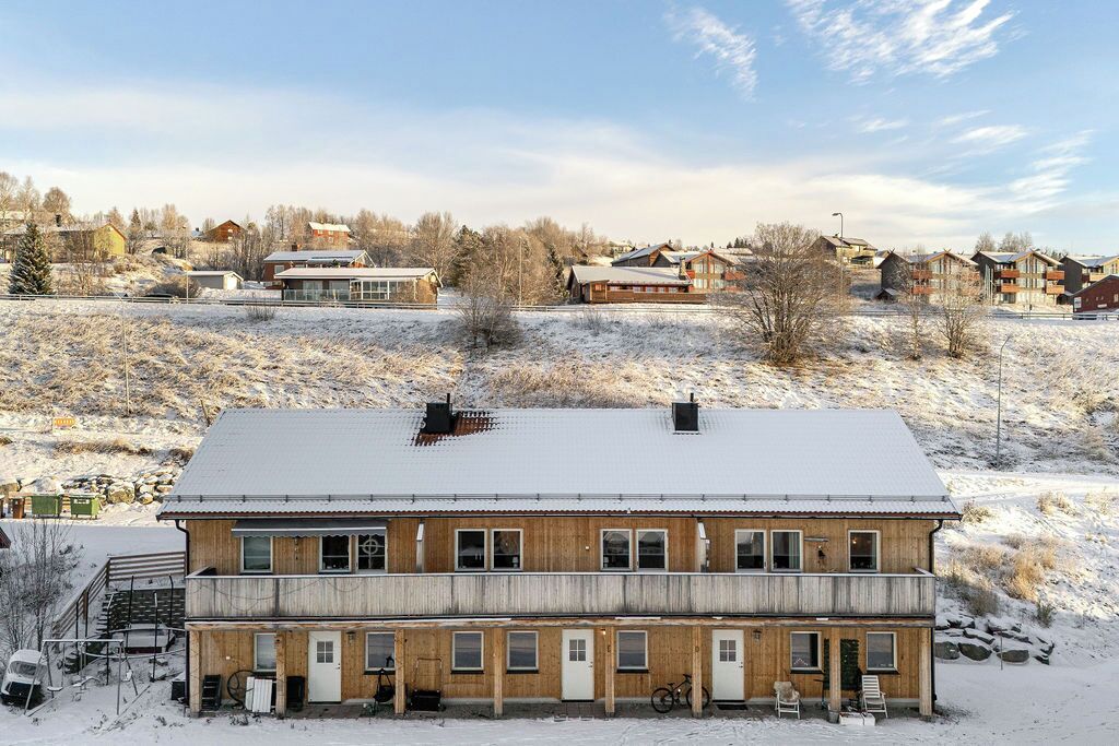 A two-story wooden building with a snow-covered roof, a front porch with steps, and a visible garage.