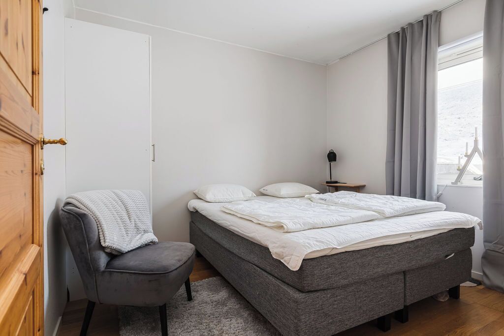 A bedroom with a bed, a chair, a bedside table, and a window with curtains.