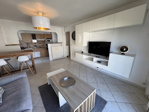 Un salón moderno con televisor de pantalla plana, una mesa de centro de madera, un sofá gris y una zona de cocina con microondas y armarios.