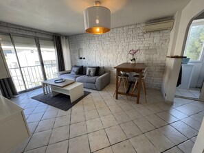 A modern living room with a grey sofa, a wooden coffee table, a dining table with chairs, and a large window with a view. - Apartment “Les Floralies” with Shared Garden, Wi-Fi, and Air Conditioning (La Garde)