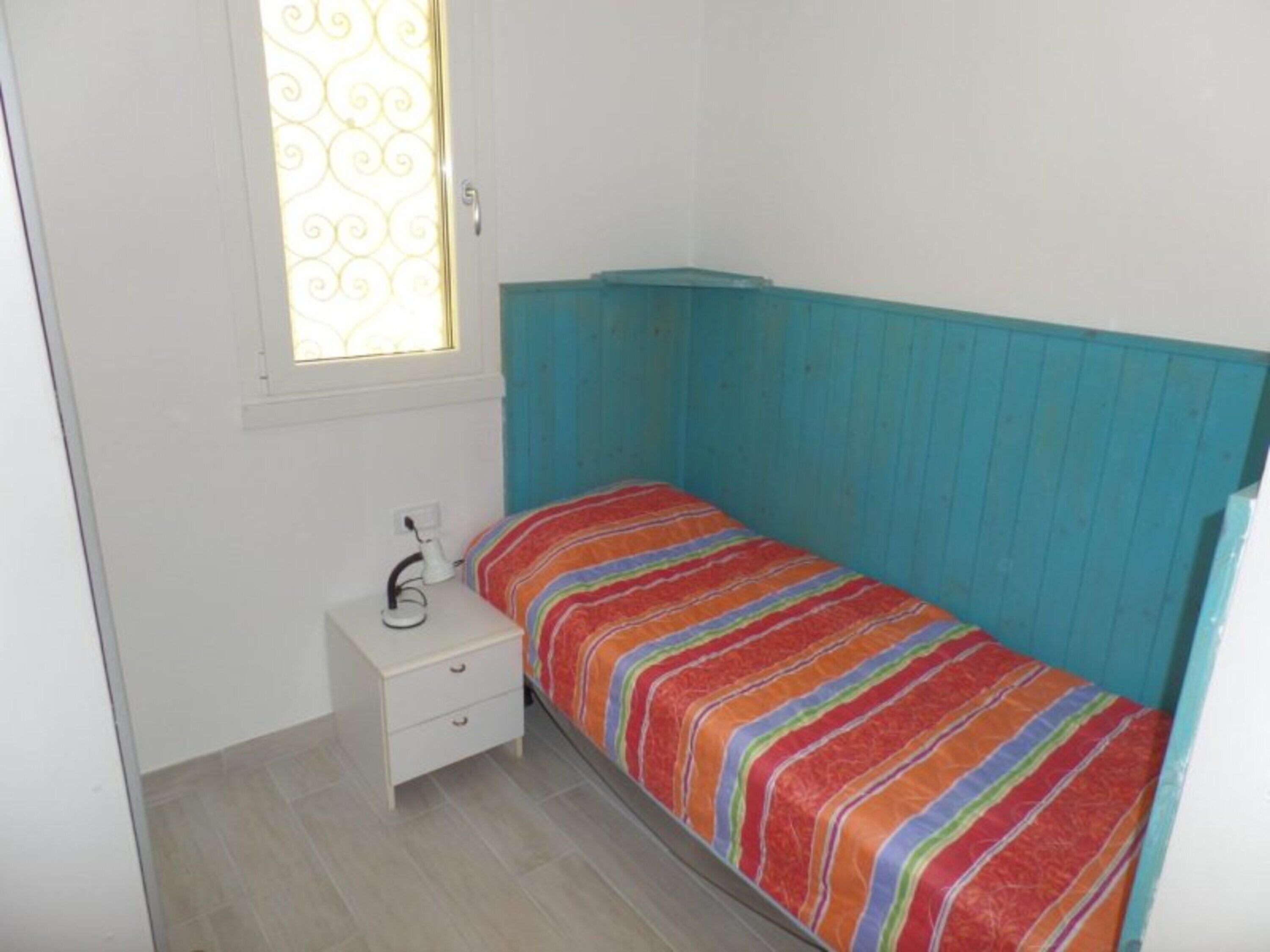 Une petite chambre avec un lit simple, une table de chevet blanche, une fenêtre ornée d’un motif décoratif et une tête de lit bleue.