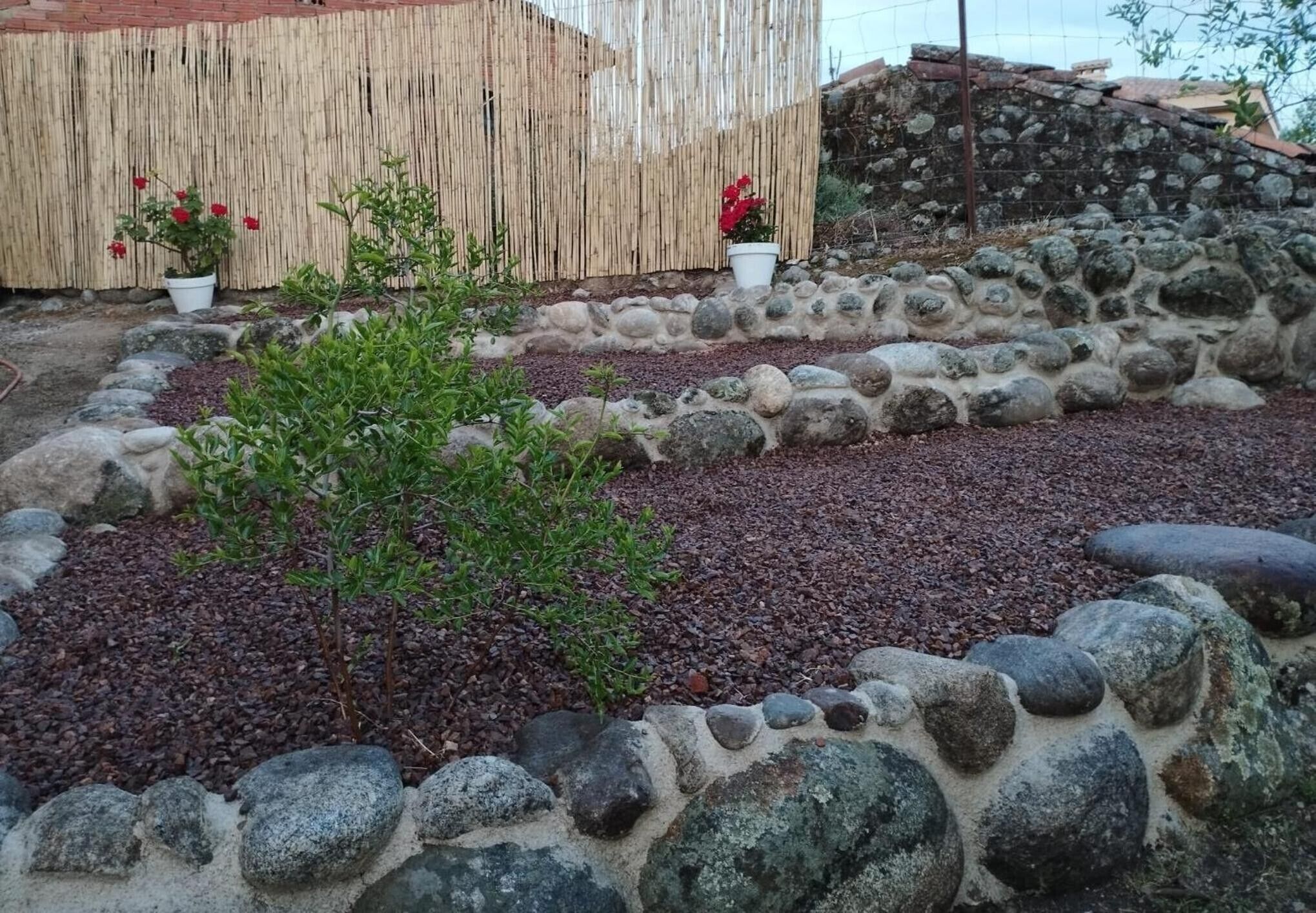 Un jardin avec un mur en pierre, des plantes en pot et une clôture en bambou.