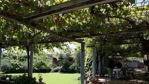 Een overdekt terras met een tafel en stoelen, omgeven door groen en uitzicht op een gazon.