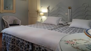 Een net opgemaakt bed met een bloemenbedekking, een witte stoel, een nachtkastje met een lamp en een spiegel aan de muur.