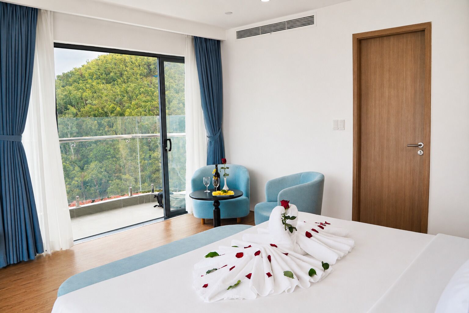 Ein Hotelzimmer mit einem Bett, einem blauen Sessel, einer Holztür und einem Balkon mit Blick auf Grünflächen.