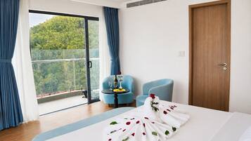 Ein Hotelzimmer mit einem Bett, einem blauen Sessel, einer Holztür und einem Balkon mit Blick auf Grünflächen.