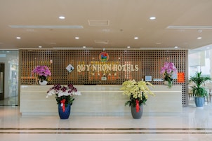 A hotel reception desk with a wooden backdrop, a sign reading 'Ouy Non Hotels,' and two large flower arrangements.